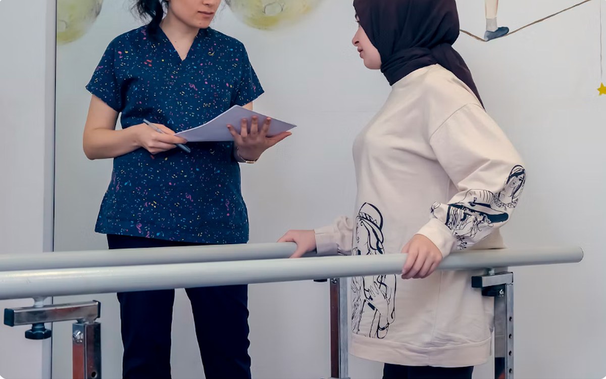 A therapist reviewing notes while a patient walks between parallel bars during gait training.
